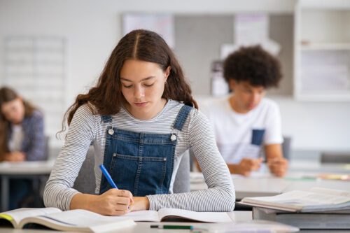 High School Girl in overalls sitting at her desk, writing in a workbook